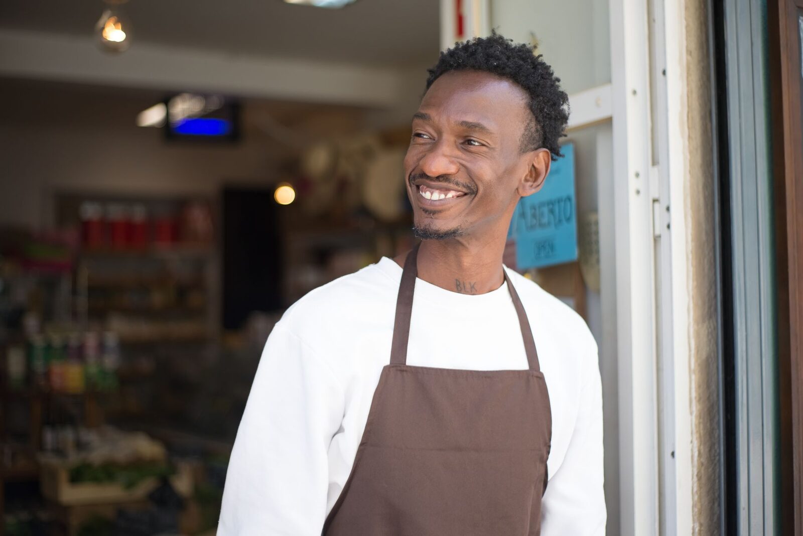 Dueño de una tienda con delantal, sonriendo en la puerta de su local